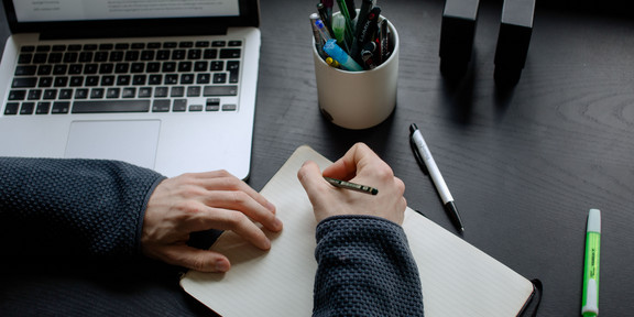 A student working at a table with a laptop, pencils and a notebook.