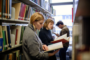 Studierende stehen lesend vor Bücherregalen in der Bibliothek.