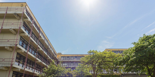 Chemiegebäude: Ein großes, breites, graues Gebäude mit Fensterfronten und Balkonen vor den Fenstern, die mit roten Metallstangen abgegrenzt werden. In der Mitte eine Treppe, auf der Menschen sitzen und links vor der Treppe Fahrräder in Fahrradständern.